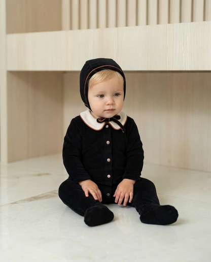 Blonde baby boy wearing Dottie Lou Classic Noir black velour footie pajama with matching hat, ivory Peter Pan collar and orange piping, sitting on a marble floor — baby model photo 2