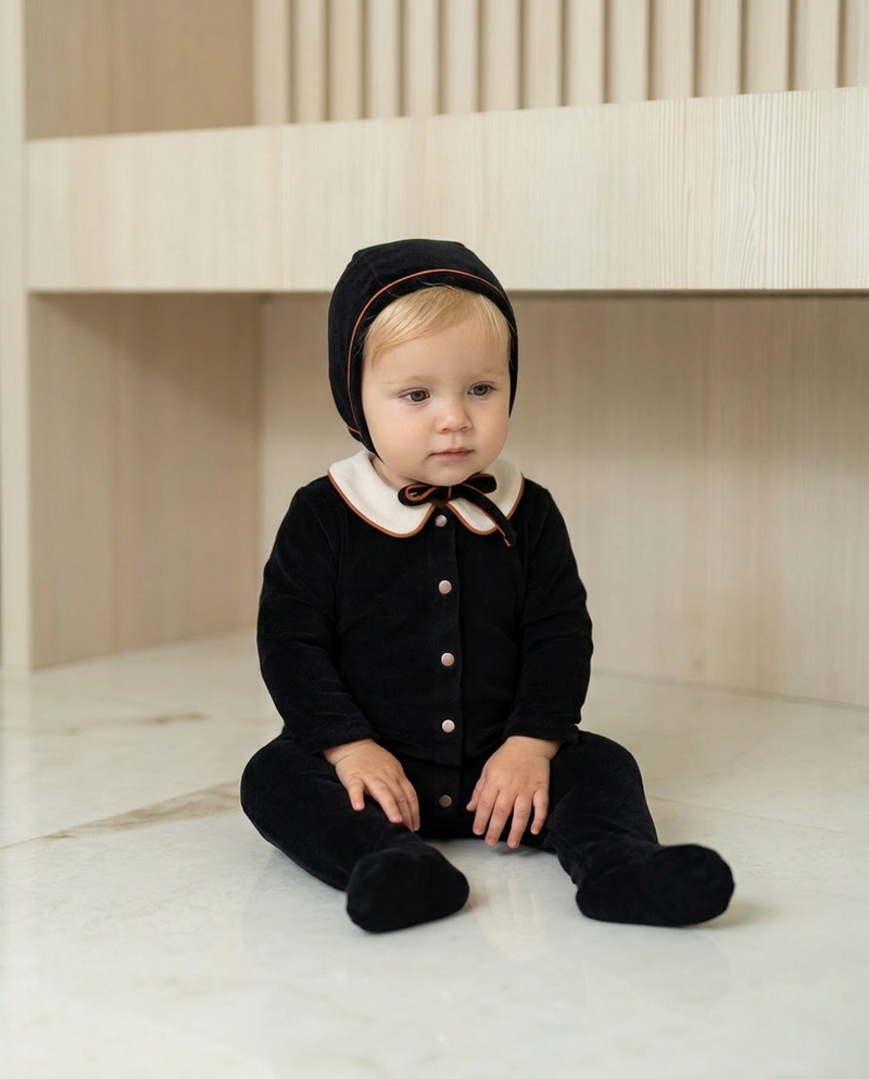 Blonde baby boy wearing Dottie Lou Classic Noir black velour footie pajama with matching hat, ivory Peter Pan collar and orange piping, sitting on a marble floor — baby model photo 2