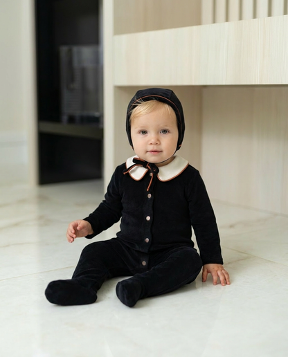 Blonde baby boy wearing Dottie Lou Classic Noir black velour footie pajama with matching hat, ivory Peter Pan collar and orange piping, sitting on a marble floor — baby model photo 1