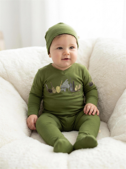 Dottie Lou Melody music-print footie pajama with matching hat in moss green worn by baby boy sitting on white sheepskin chair